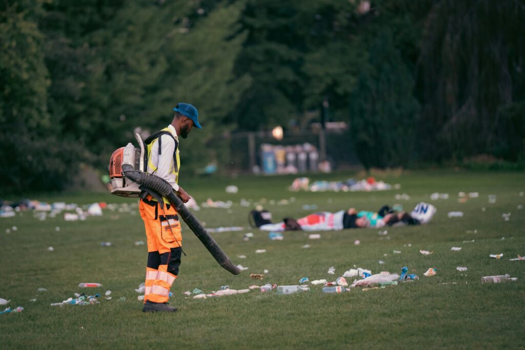 Man with leaf blower cleans littered park with people resting