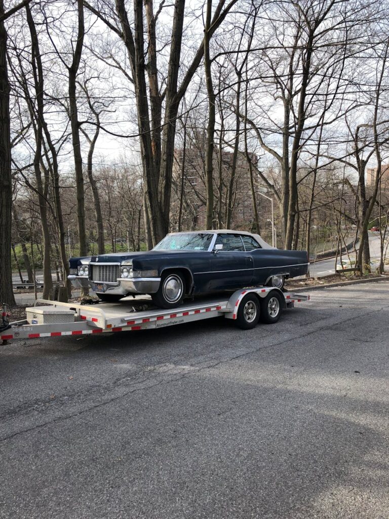 Classic blue car on a trailer amidst leafless trees on a street.