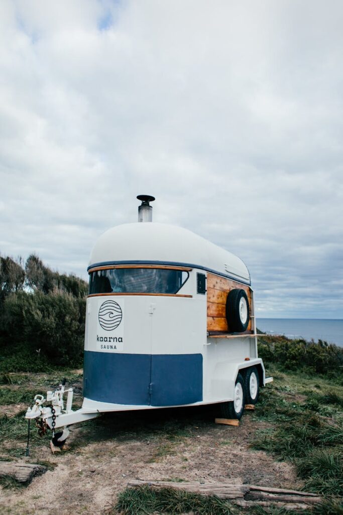 A mobile sauna trailer parked on a grassy coastline under a cloudy sky.
