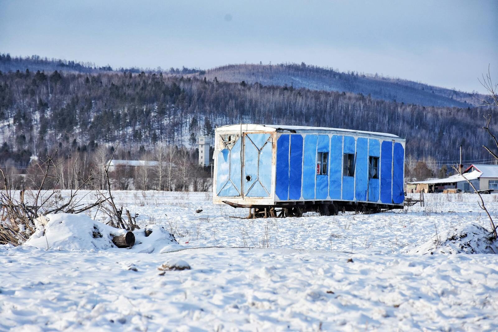 Blue trailer in snowy field with mountains in the background, capturing cold winter scenery.