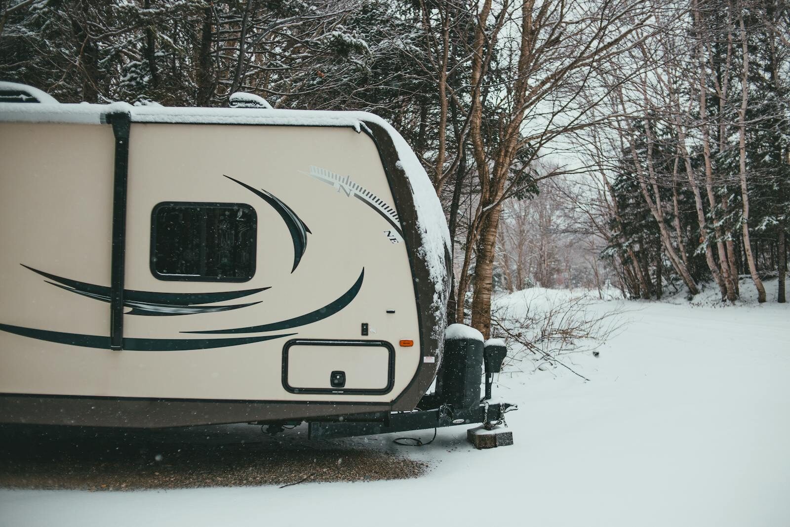 Photo by Erik Mclean Camper trailer parked in coniferous and deciduous forest on road trip in snowfall in gloomy weather in winter in daytime