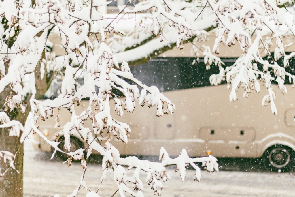 Snow falls on tree branches near a recreational vehicle.