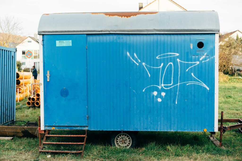 Blue construction trailer with graffiti on a grassy field, surrounded by pipes.