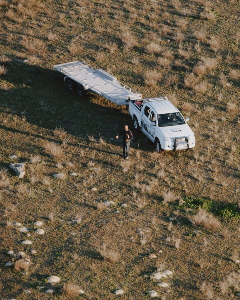 Overhead shot of a white truck with a trailer on a dry, grassy landscape. High contrast daylight scene.