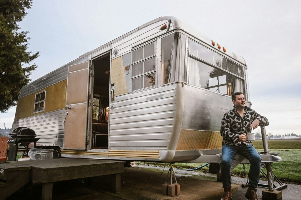 A man sitting beside a vintage travel trailer outdoors.